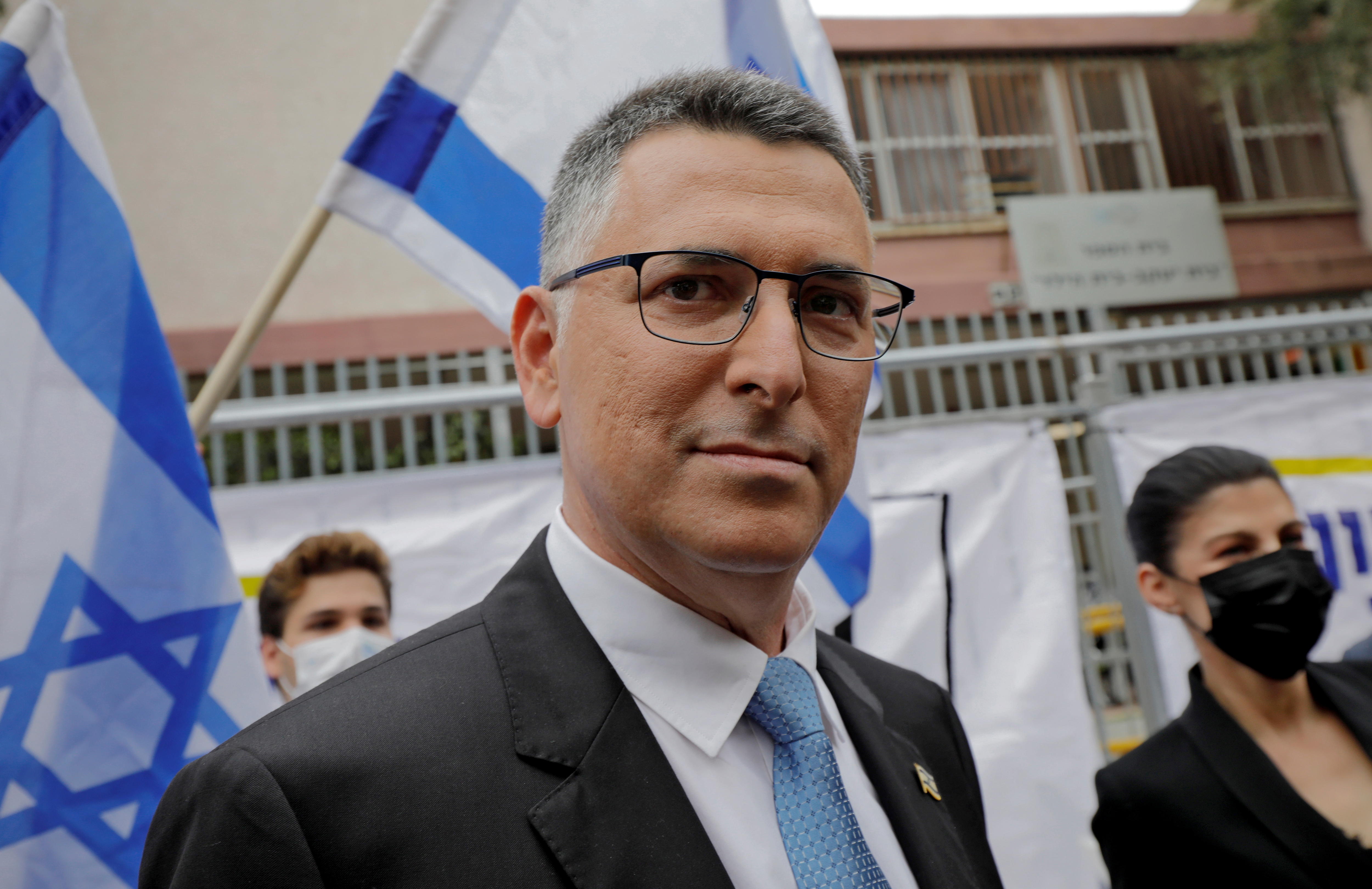 man wearing glasses and black suit mid shot with blue and white flags in background
