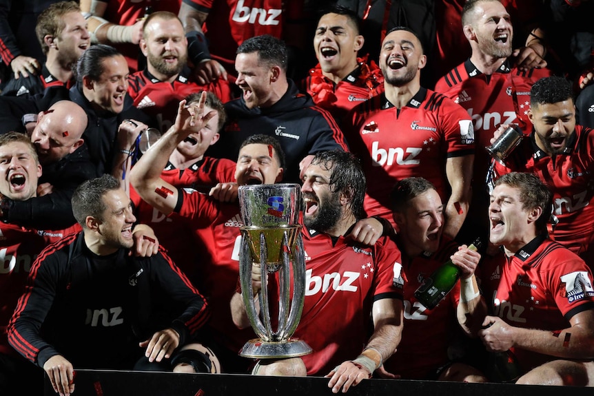 Male rugby union players sing and smile as they sit and stand with a trophy in front of photographers.