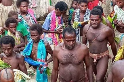 Two naked men from Vanuatu wear traditional genital coverings while walking in front of a group of women in grass skirts.