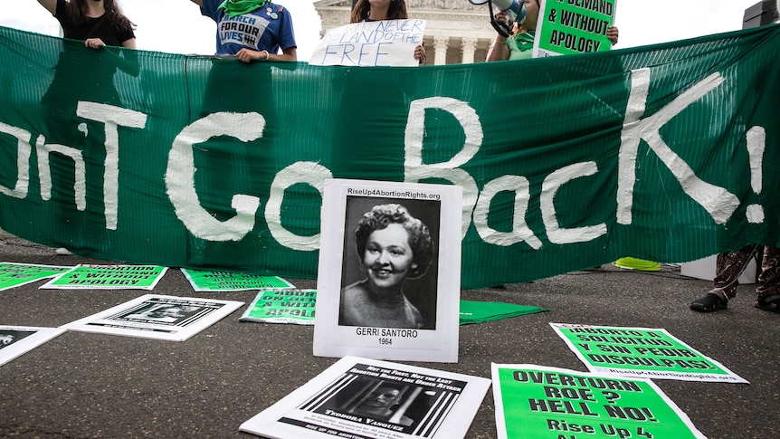 A photo of Gerri Santoro propped up before a protest banner reading "Won't Go Back!" 