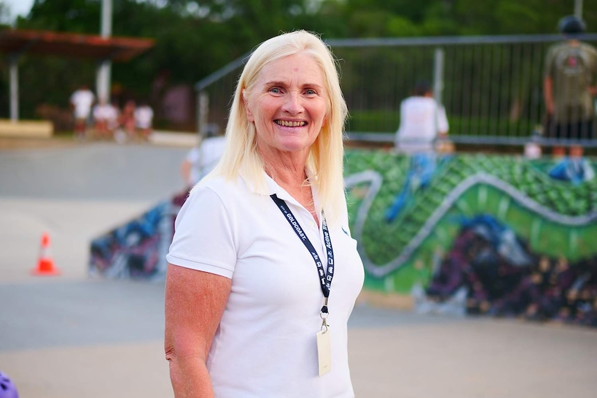 Woman in a white shirt with a lanyad and white hair standing in front of a blurred skateboard park.