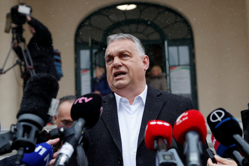 Viktor Orban, wearing a black winter coat over a sharp white shirt, frowns as he speaks, surrounded by microphones