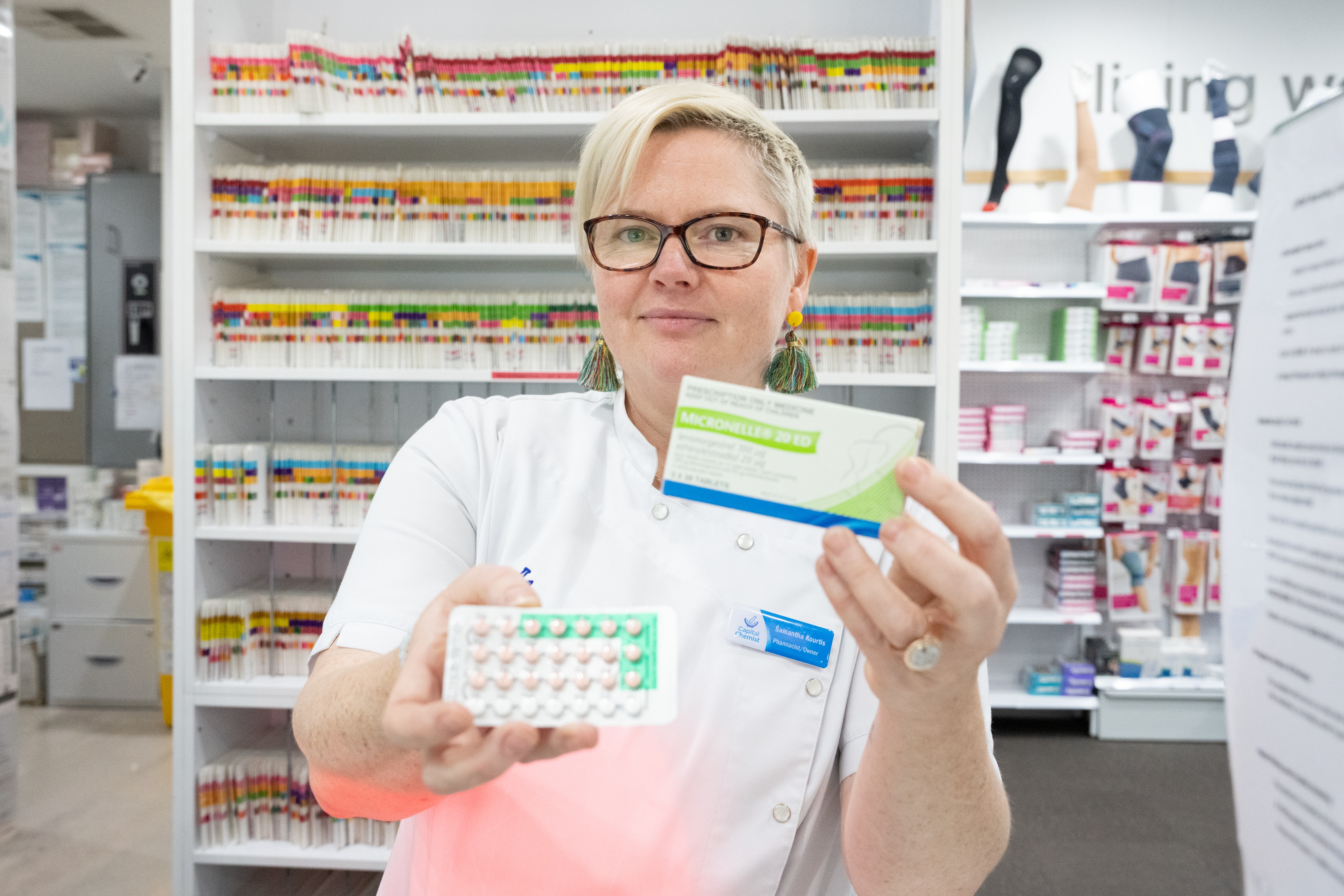 A pharmacist holds a medication box in one hand and a sheet of tablets in the other.