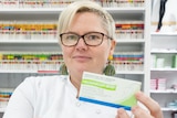 A pharmacist holds a medication box in one hand and a sheet of tablets in the other.