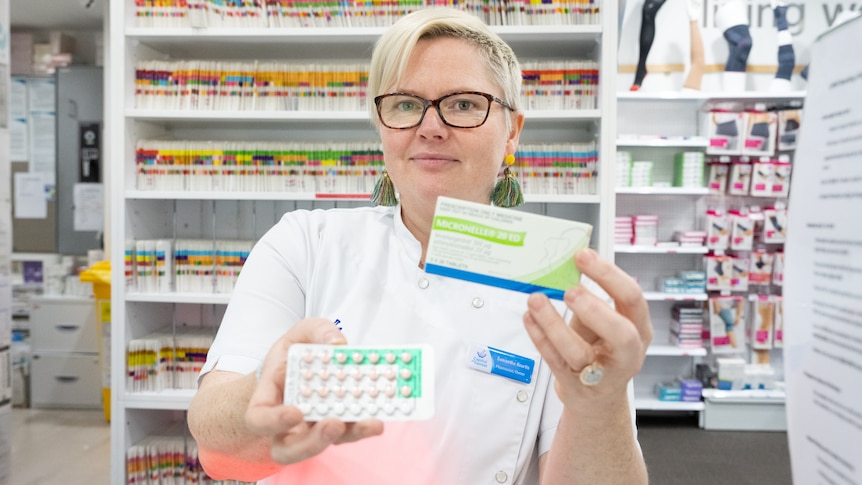 A pharmacist holds a medication box in one hand and a sheet of tablets in the other.