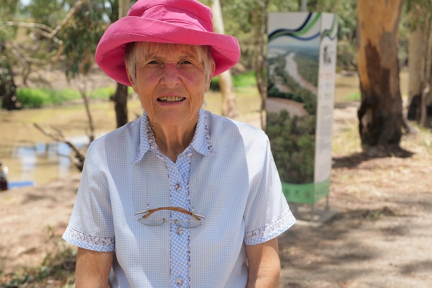 An older, silver-haired woman smiling, wearing pink hat, blue and white button-up shirt with glasses hanging from her neck.