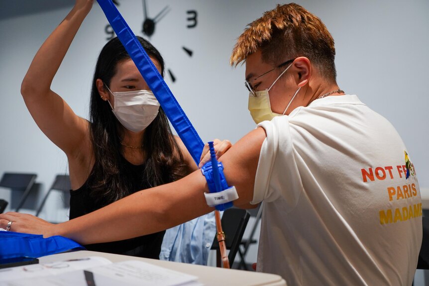 A woman applies a blue tourniquet bandage with tension strap to the arm of a man, both wear masks