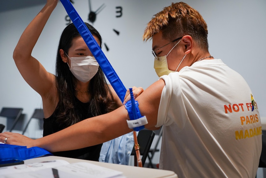 A woman applies a blue tourniquet bandage with tension strap to the arm of a man, both wear masks