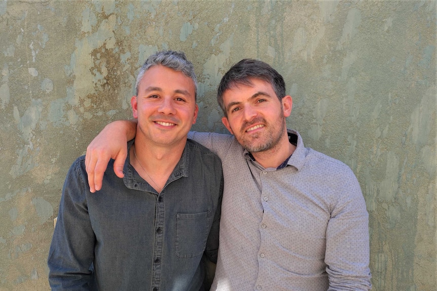 Two men against a wall smile at the camera, with their arms around each other.