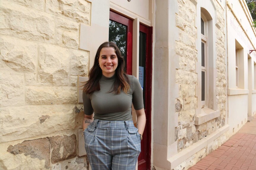 A young woman stands with her hands in her pockets smiling at the camera in front of a brick building