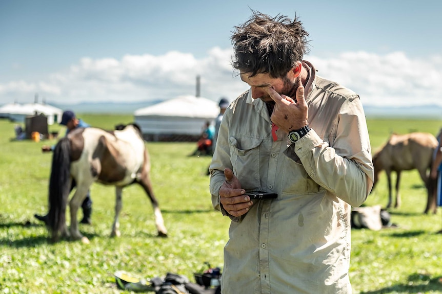 A man wipes his face while holding a phone in a paddock surrounded by horses.