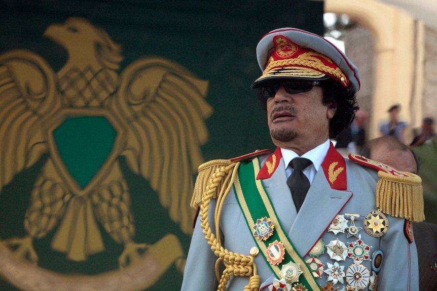 A Libyan man in military uniform, with many large medals and a hat, standing in front of a bird insignia
