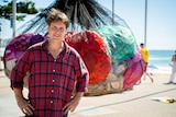 Man standing in front of giant net carrying large, different coloured plastic bags, stuffed with smaller bags on beach.