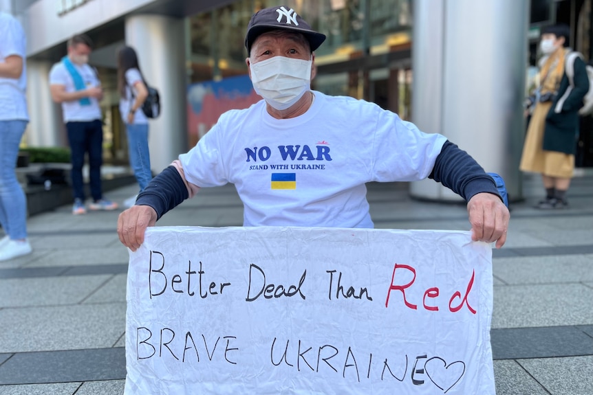 An older man in a NO WAR T-shirt holds a banner saying "Better dead than red. Brave Ukraine"