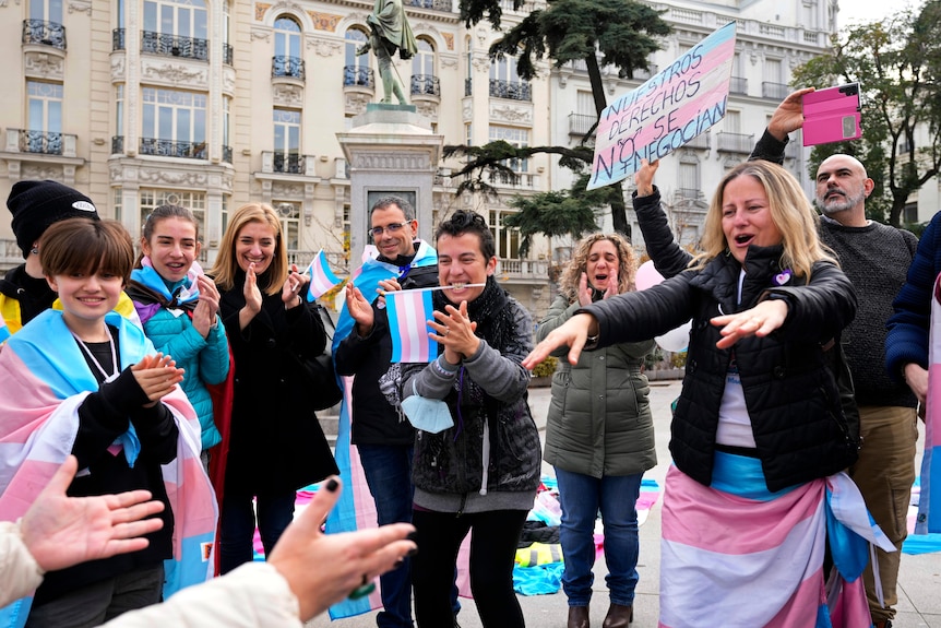 A group of people clapping holding pink blue and white flags