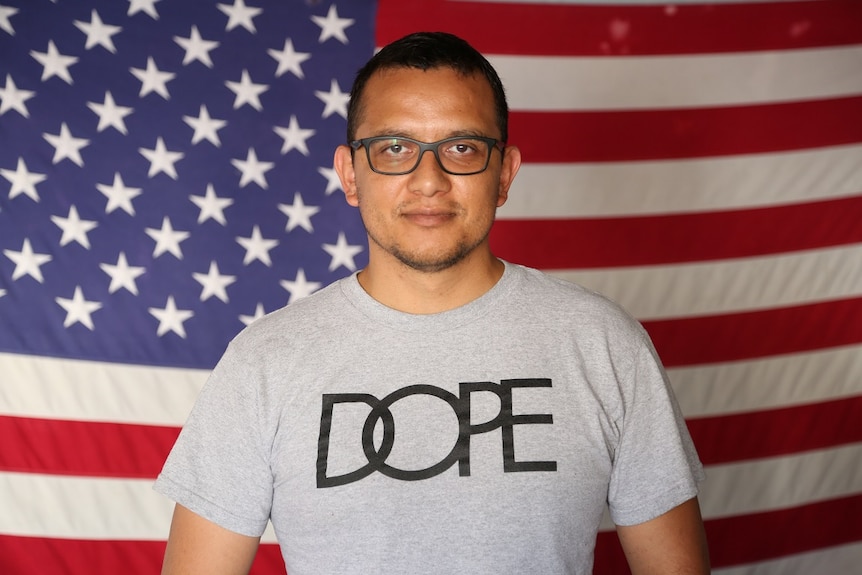 Man wearing t-shirt and glasses stands in front of US flag.