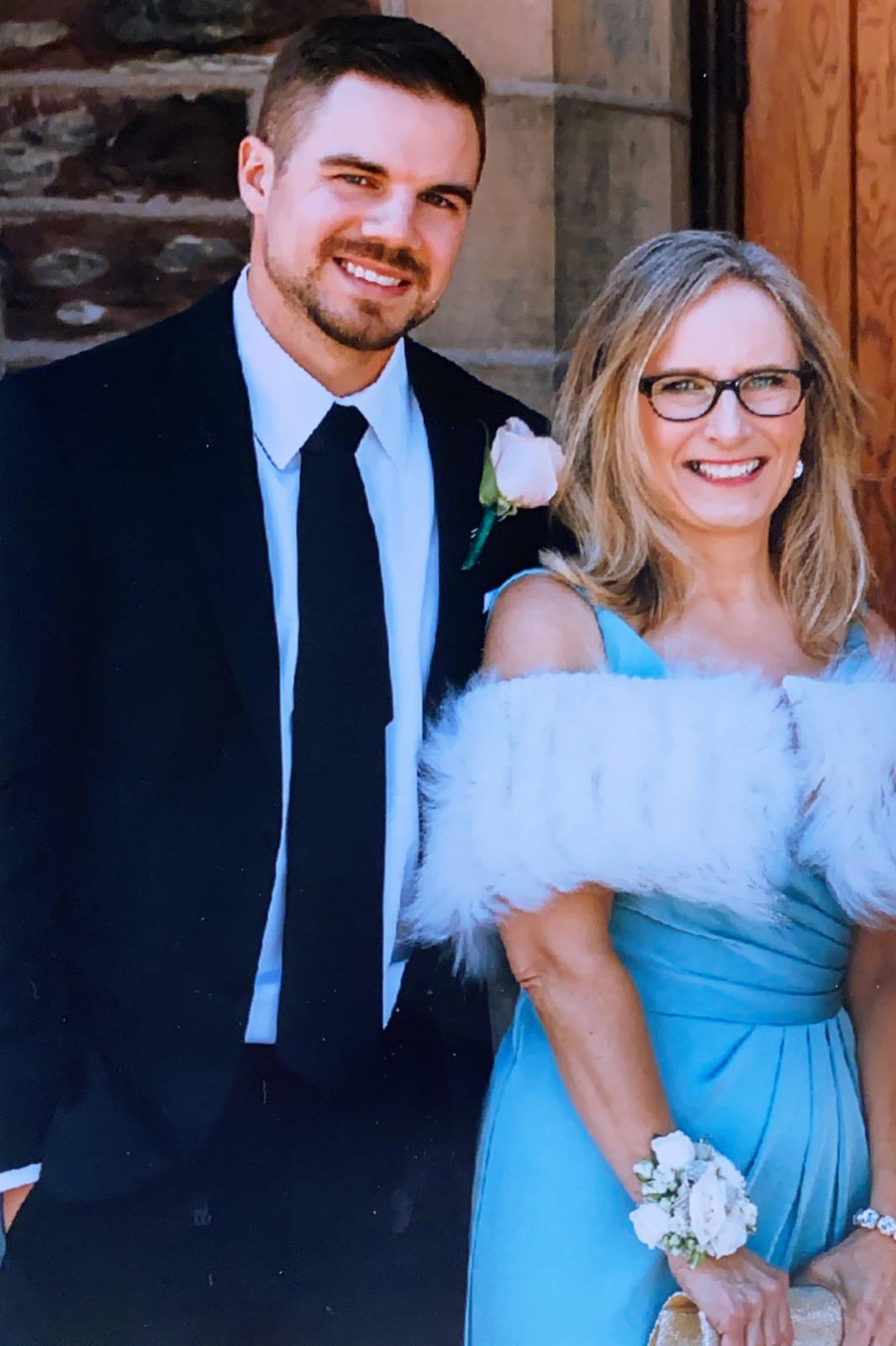 Marc smiles wearing a black suit with a flower in his breast pocket. Denise stands ust in front os him in a formal blue dress. 