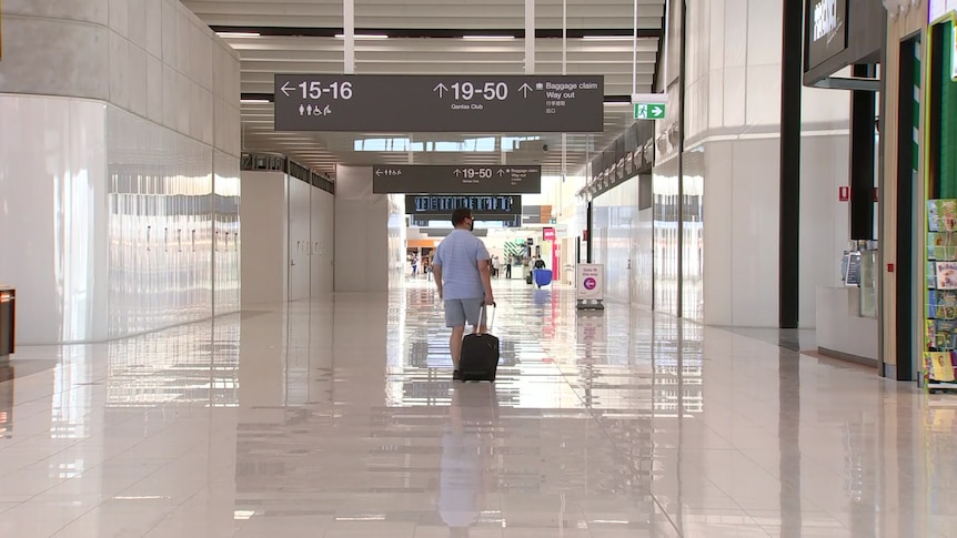 A man wearing shorts and a shirt pulls a suitcase through an empty airport corridor
