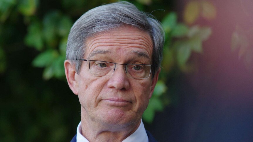 A close up photo of Mike Nahan standing in front of green bushes.