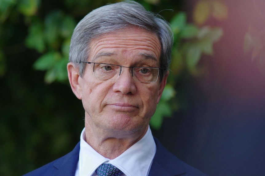 A close up photo of Mike Nahan standing in front of green bushes.