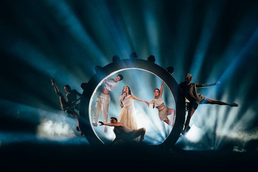 Eden Golan with mic and arms out, white dress surrounded by dancers in a circle