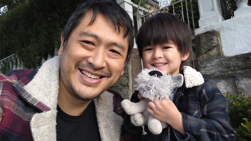 A father and his preschool-aged son smile happy. The boy is cuddling a soft toy