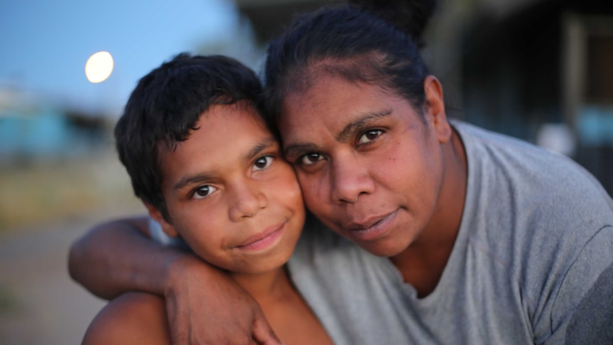 An Aboriginal woman leans close and hugs an Aboriginal boy.
