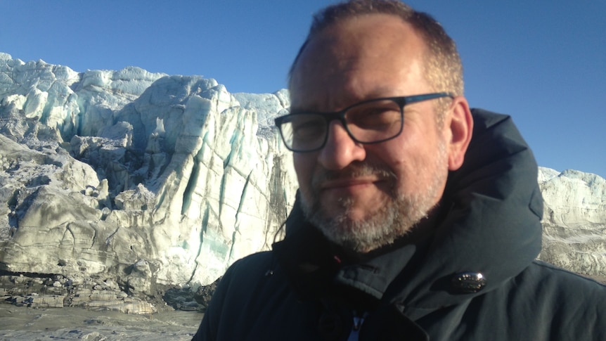 Head shot of man wearing glasses in puffer jacket with glacier in background.