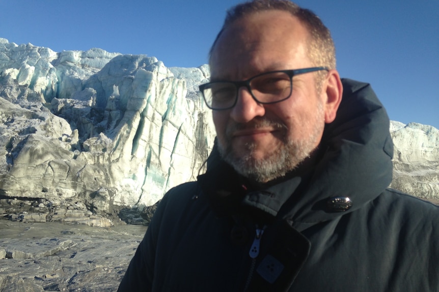 Head shot of man wearing glasses in puffer jacket with glacier in background.