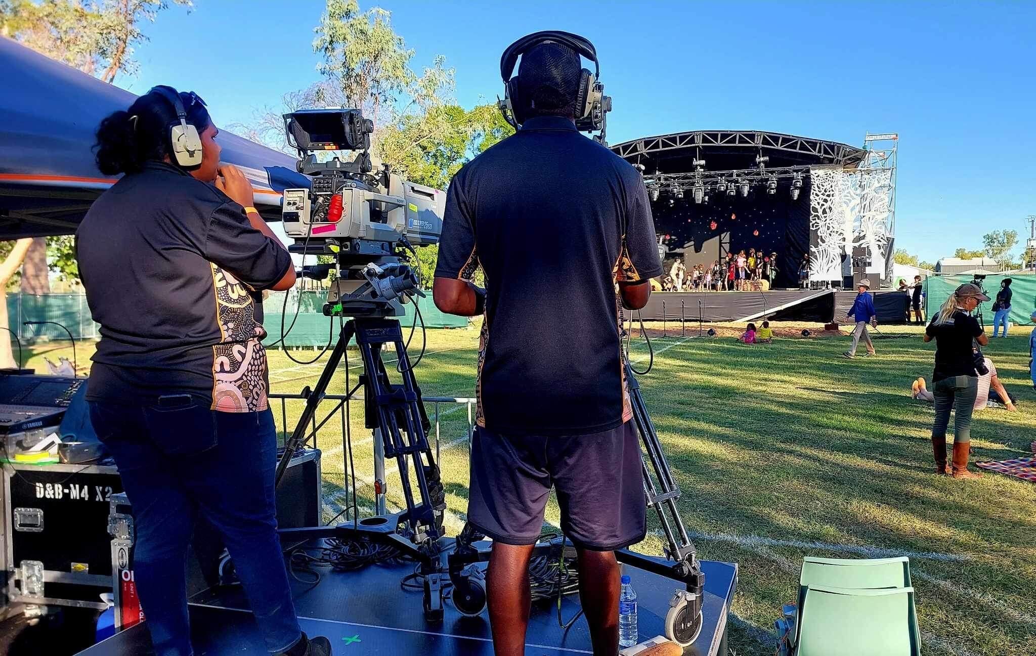 Camera operators and cameras on tripods filming an outdoor stage in a park.