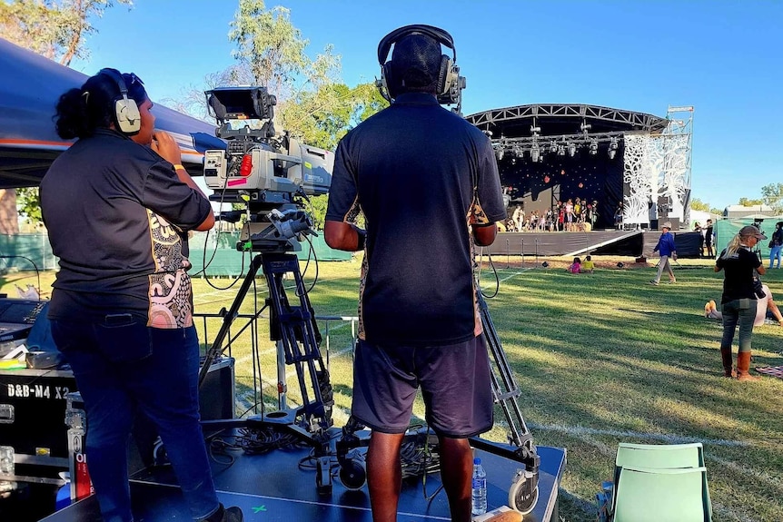 Camera operators and cameras on tripods filming an outdoor stage in a park.