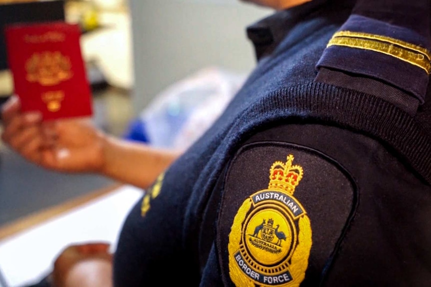 An Australian Border Force officer, with badge in foreground, holds a passport in background.