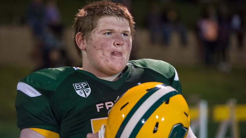 Eddie Sullivan looks his lips in mid-shot. He wears a football jersey and holds a helmet in his hands. He looks away.