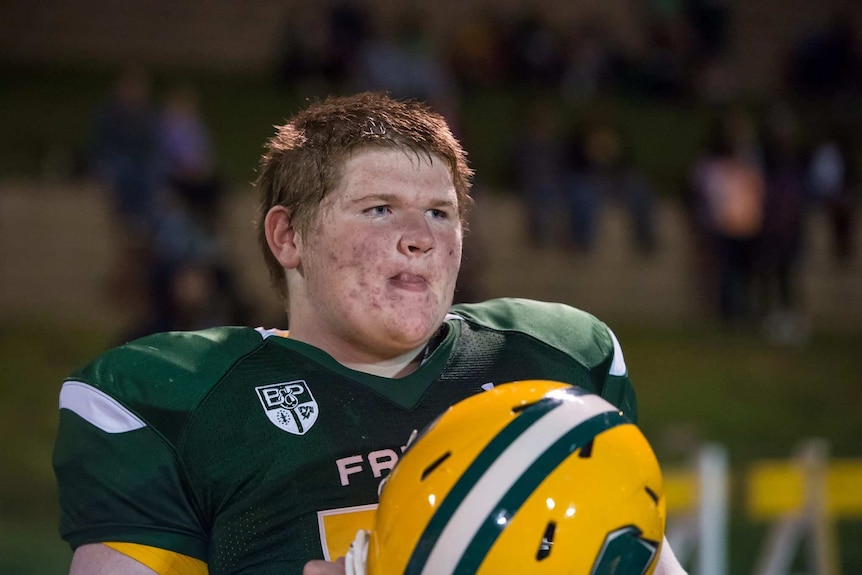 Eddie Sullivan looks his lips in mid-shot. He wears a football jersey and holds a helmet in his hands. He looks away.