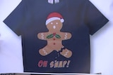 A picture of a t-shirt with a gingerbread man on it wearing a Santa hat, on a white background.