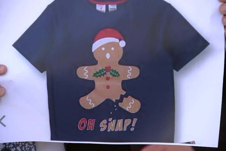 A picture of a t-shirt with a gingerbread man on it wearing a Santa hat, on a white background.