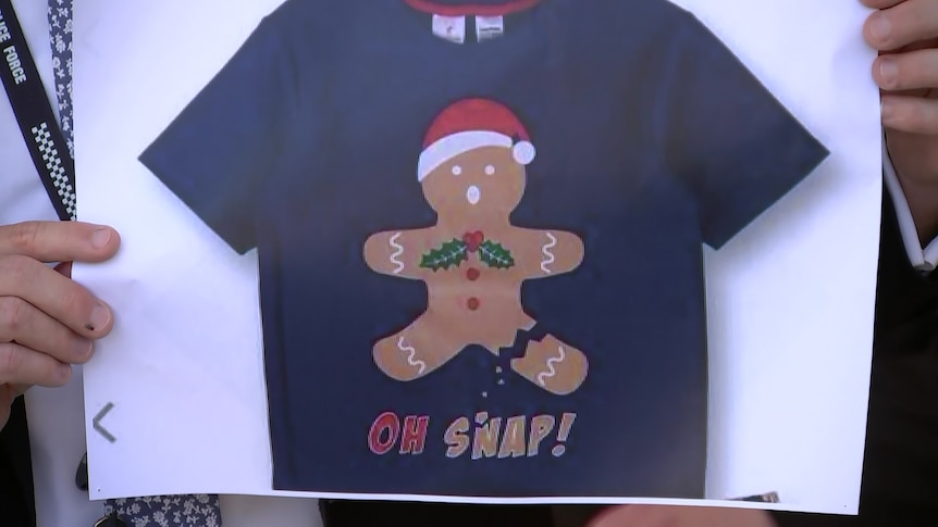 A picture of a t-shirt with a gingerbread man on it wearing a Santa hat, on a white background.