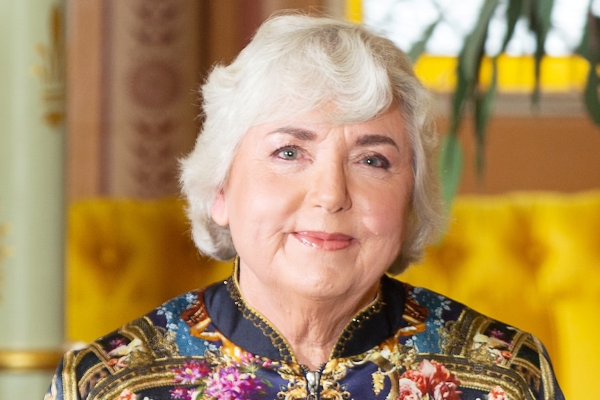 A woman with white hair and a colourful top smiles.