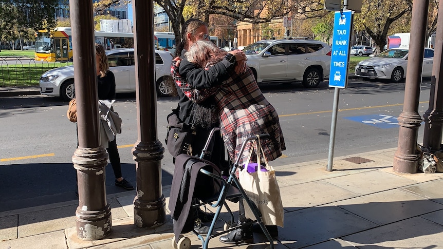 A woman with a walker is hugged by a man on a city footpath