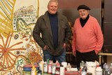 An elderly man and his adults son stand in an art studio in front of canvas