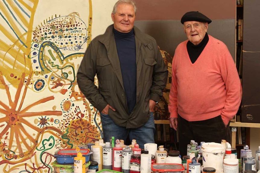 An elderly man and his adults son stand in an art studio in front of canvas