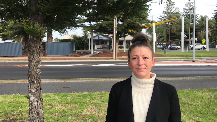 A woman wearing a cream jumper and black blazer with her hair in a bun stands in front of a caravan park