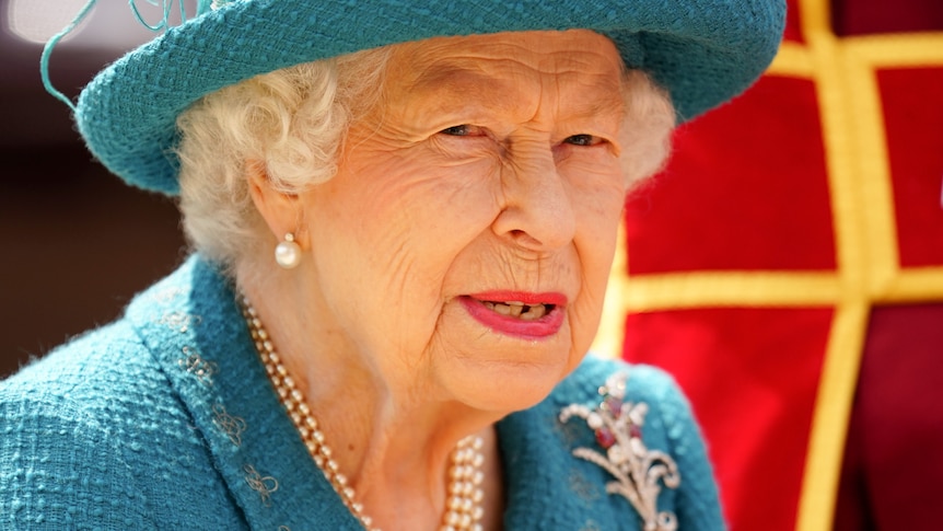 Queen Elizabeth II wears a blue coat with matching hat and pearl necklace as she looks away from the camera.