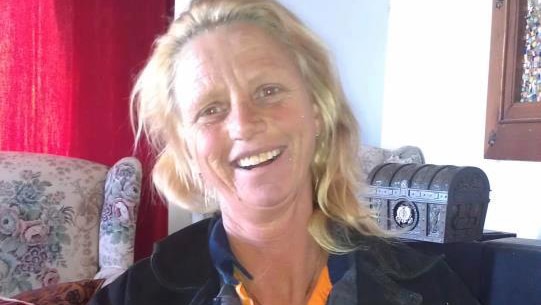 Julie Cooper smiles posing for a photo sitting indoors wearing a black jacket, grey jumper and yellow shirt.