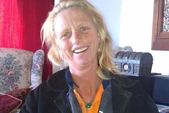 Julie Cooper smiles posing for a photo sitting indoors wearing a black jacket, grey jumper and yellow shirt.
