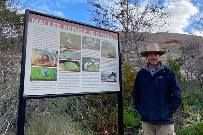 Man with hat standing in from of large rock and bush, and next to sign about Mallee Refuge Red Trail with pictures of plants