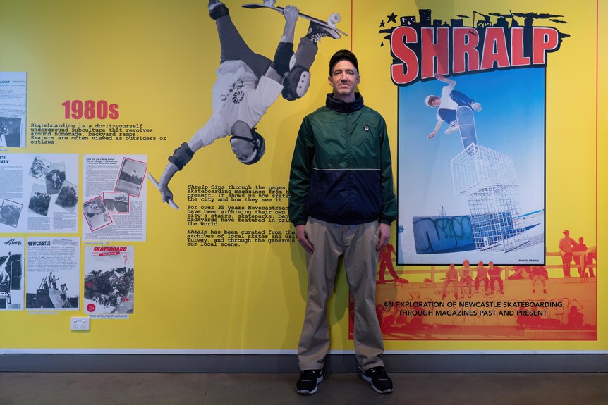 A man wearing a green jacket stands in front of a yellow wall covered in old magazine covers of people skating