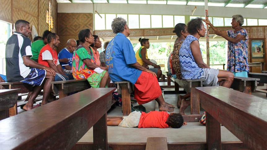 A boy lies on the ground as the adults around him, sitting in church pews, sing in a choir.