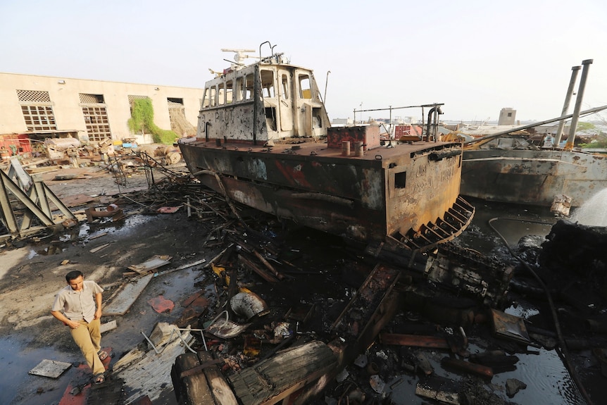A damaged tugboat covered in black marks like from fire or dirt, sits on a pile of bent amid debris and wreckage next to water