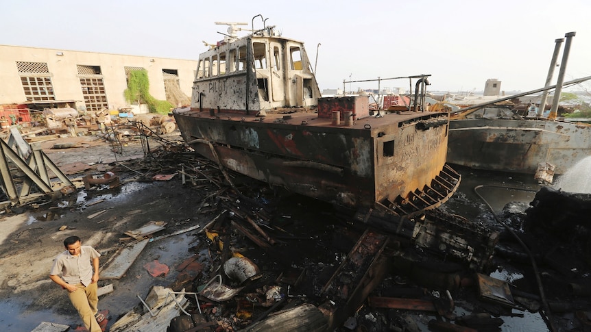 A damaged tugboat covered in black marks like from fire or dirt, sits on a pile of bent amid debris and wreckage next to water
